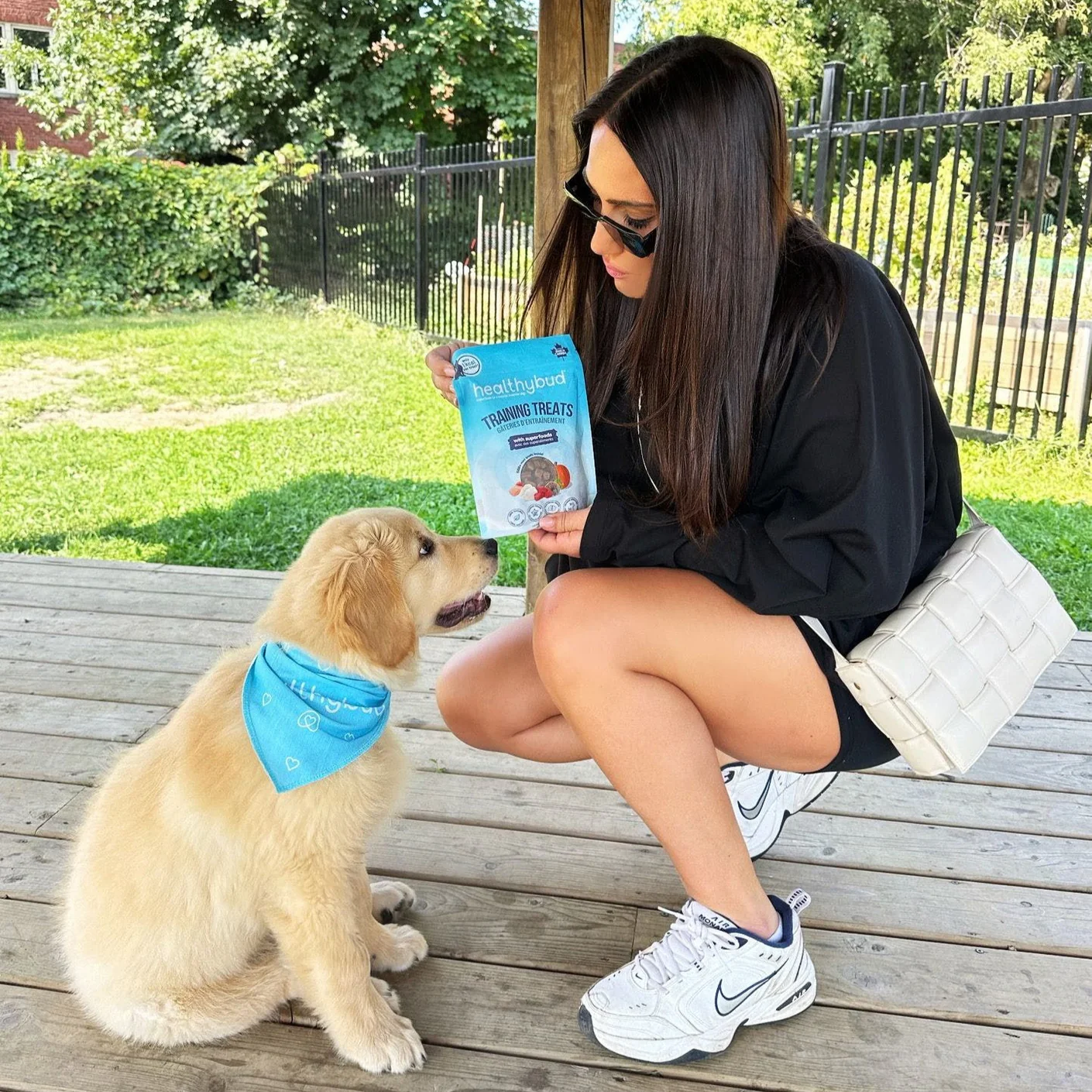 A woman crouches on a wooden deck, showing a bag of treats to a golden retriever puppy.