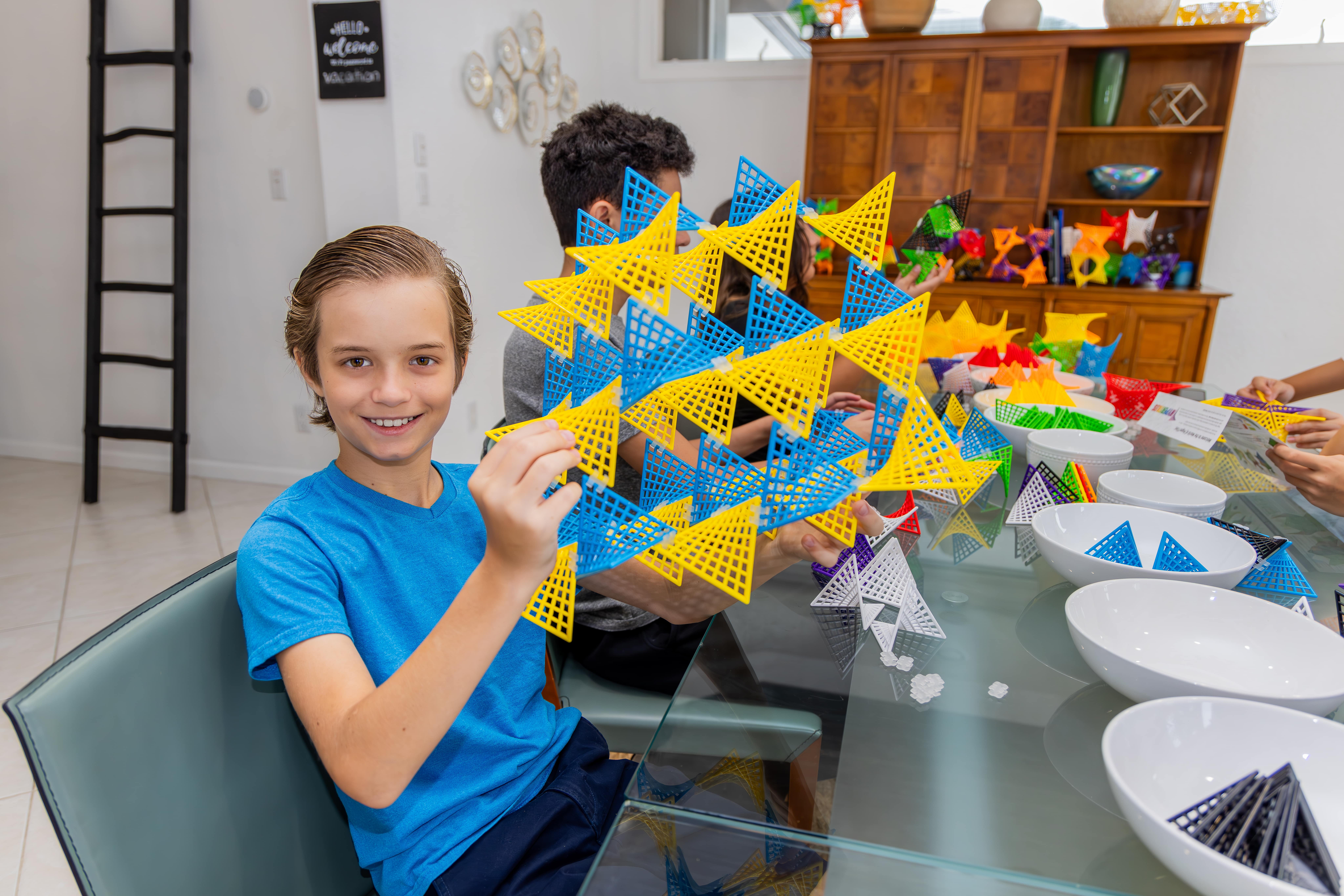 Child holding a triangular patterned structure, sitting at a glass table with more geometric shapes.