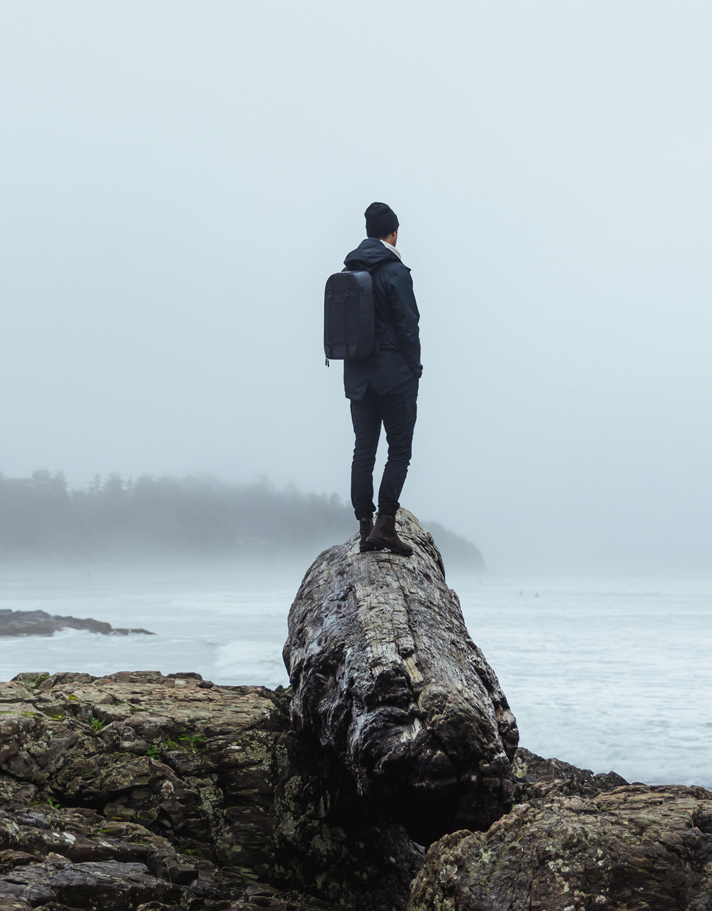 Person standing on a large rock by the ocean, wearing a backpack.