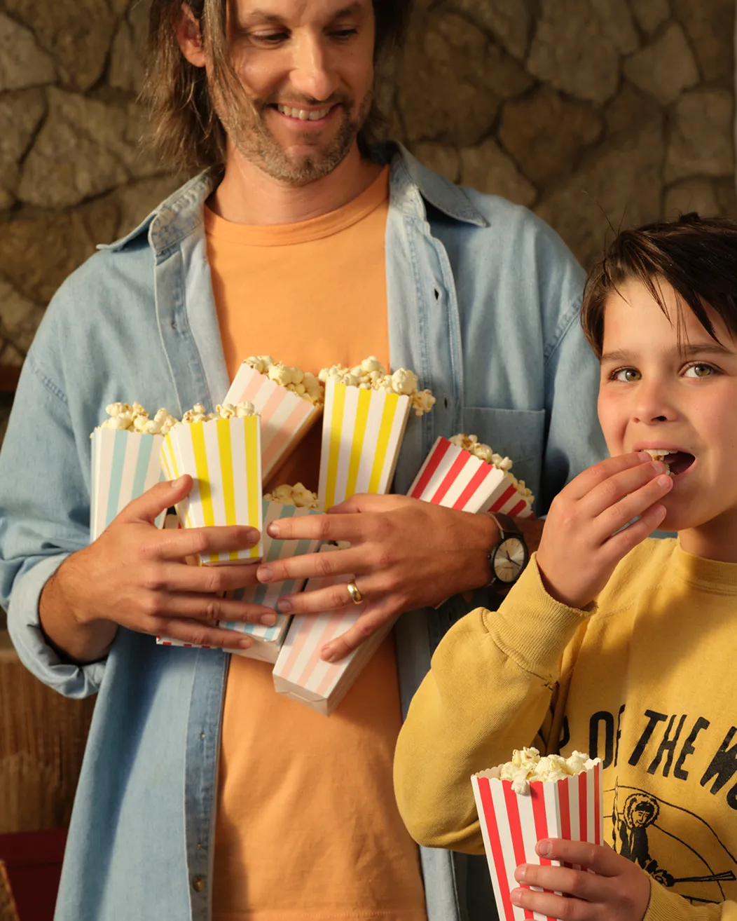 A man holds several cartons of popcorn while a child next to him eats from one.