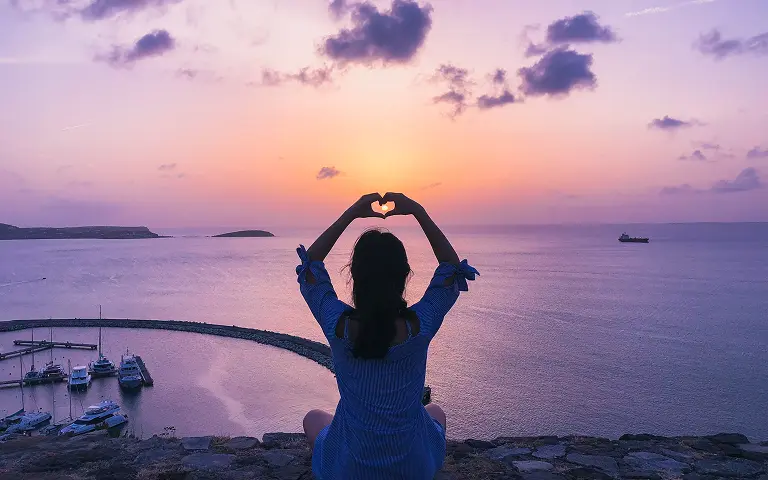 A woman sits on a cliff making a heart with her hands to frame the sunset over the sea.