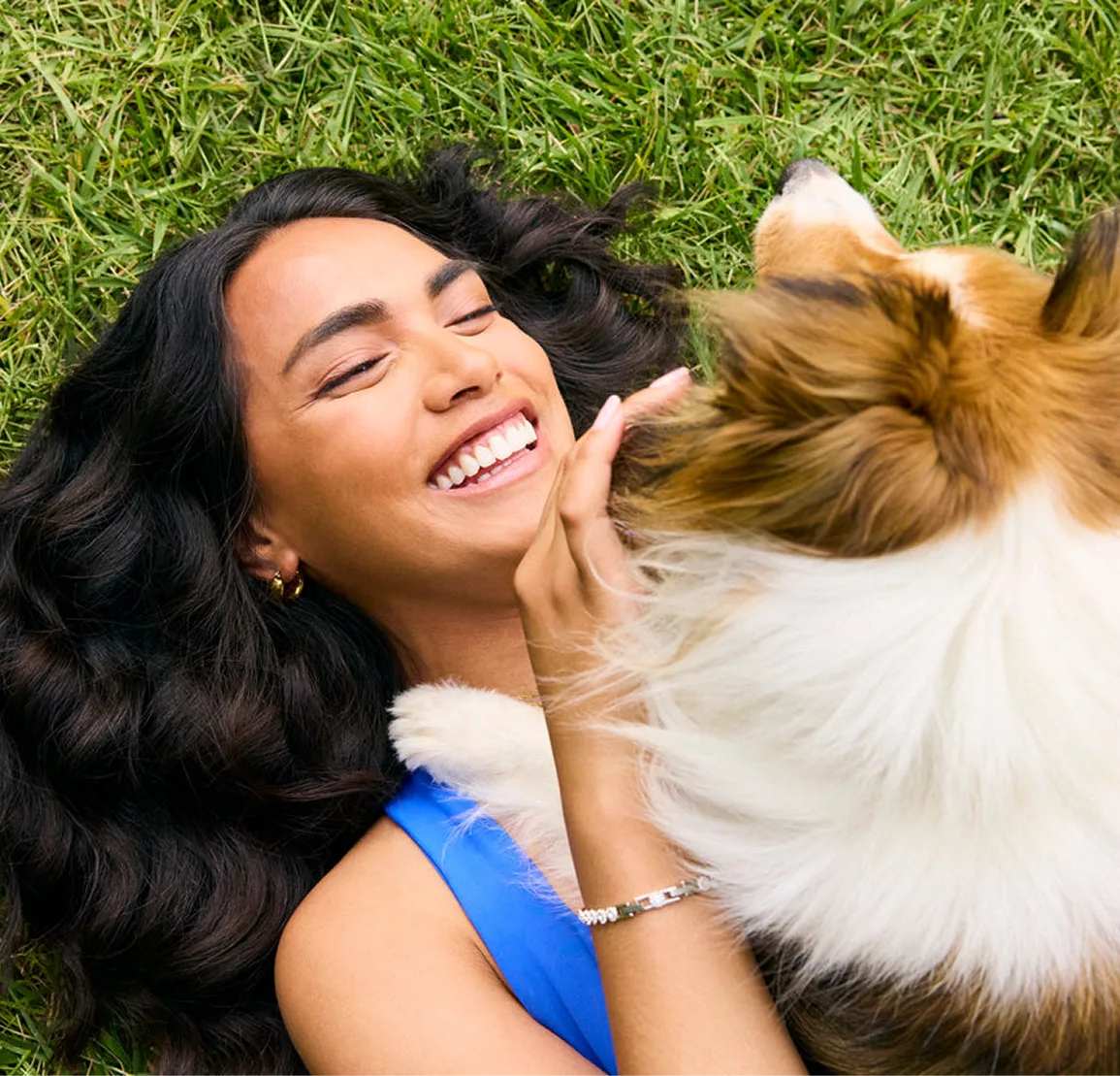 An overhead shot of a smiling woman with long dark hair lying on the grass with her dog.