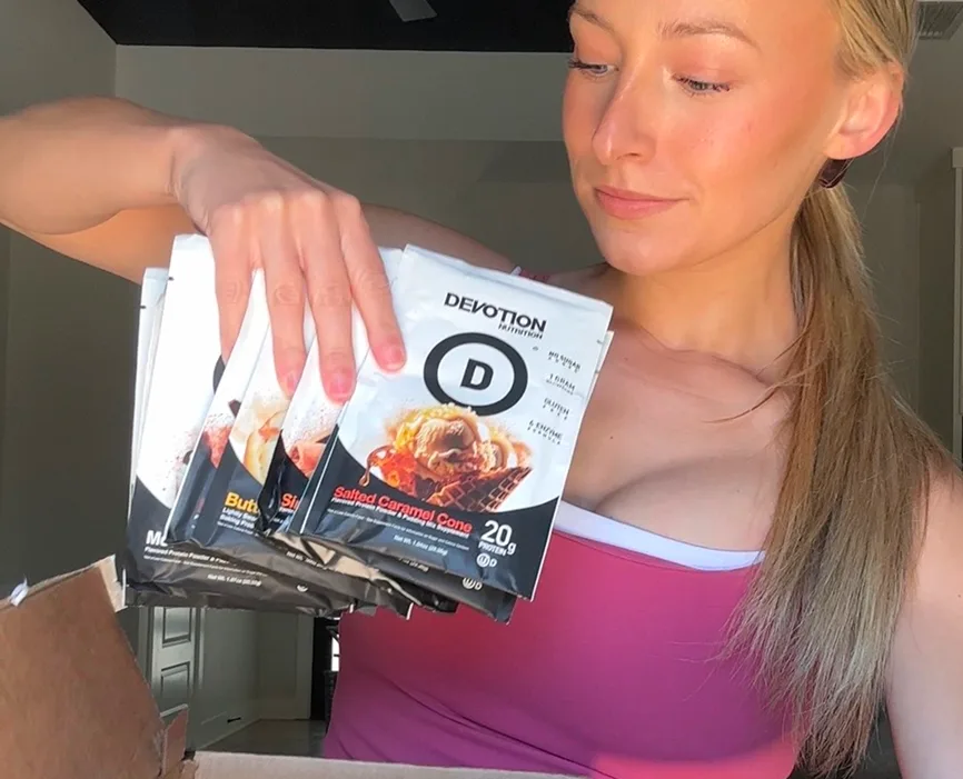 A woman holds several packets of Devotion Nutrition protein powder from a cardboard box.