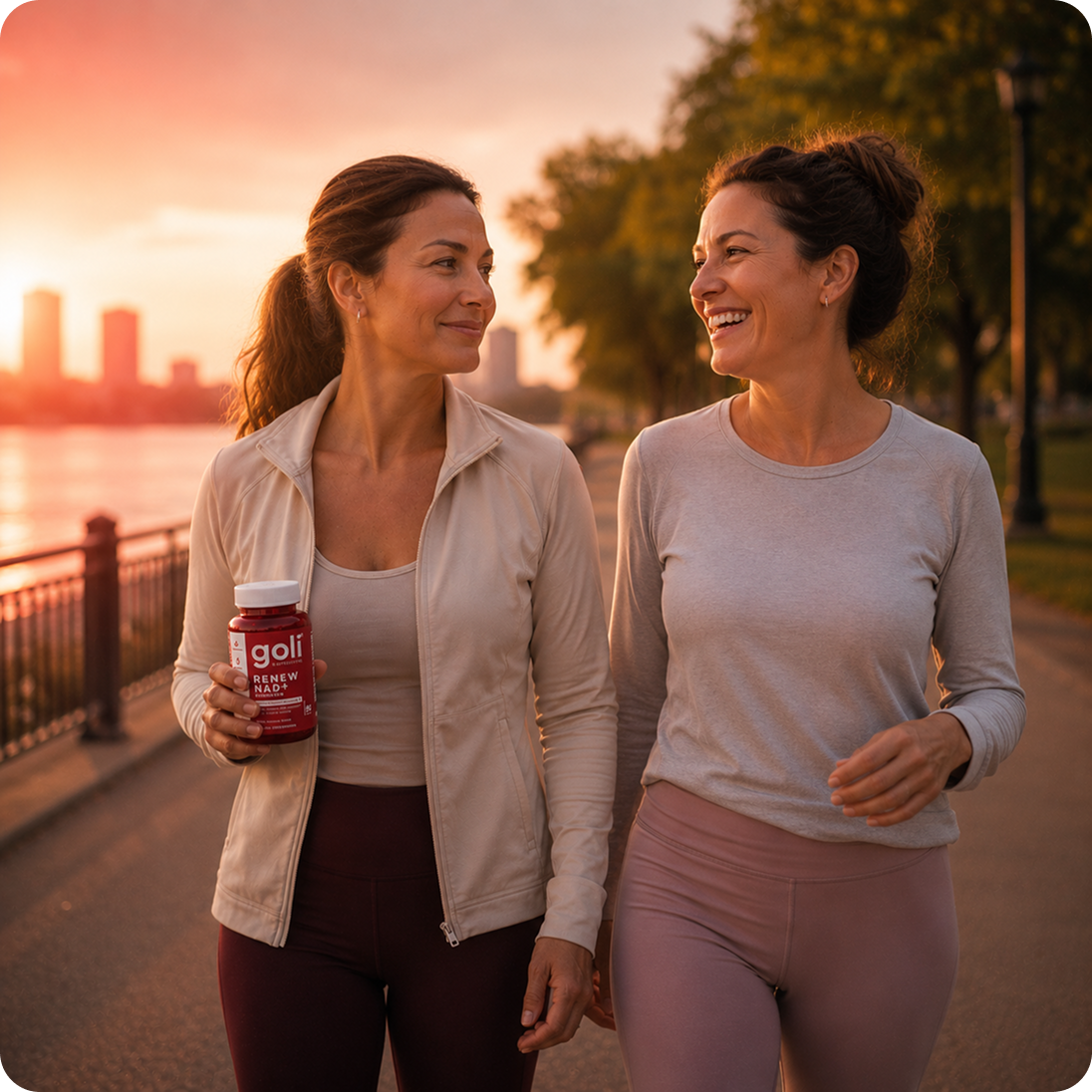 Two women in athletic wear walk along a path at sunset, one holding a red Goli bottle.
