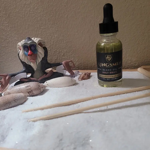 A small figurine and a bottle of beard oil on a sandy surface.