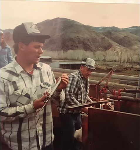Two men in plaid shirts working outdoors near a cattle chute with mountains in the background.