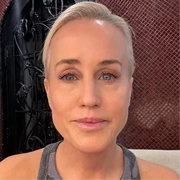 A close-up portrait of a woman with short, slicked-back blonde hair and blue eyes.