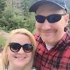 A smiling couple wearing sunglasses outdoors in a green, wooded area.