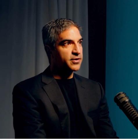 A man with salt-and-pepper hair, wearing a black jacket, speaks into a microphone.