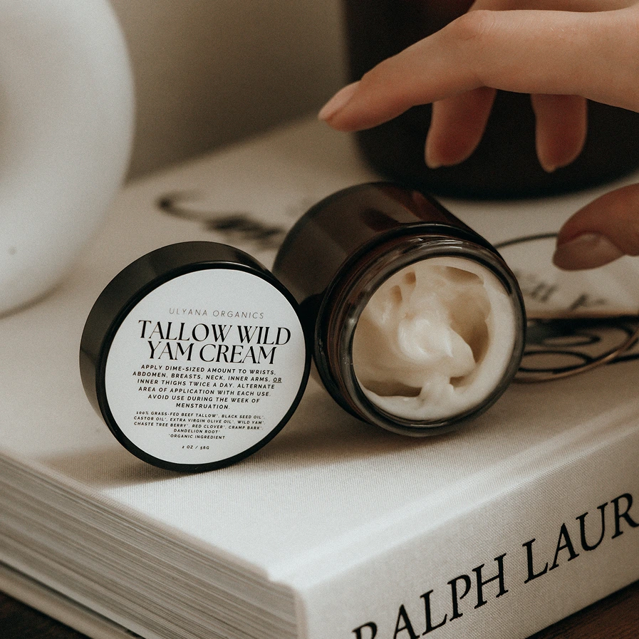 Jar of cream labeled 'Tallow Wild Yam Cream' on a book with a hand reaching for it.