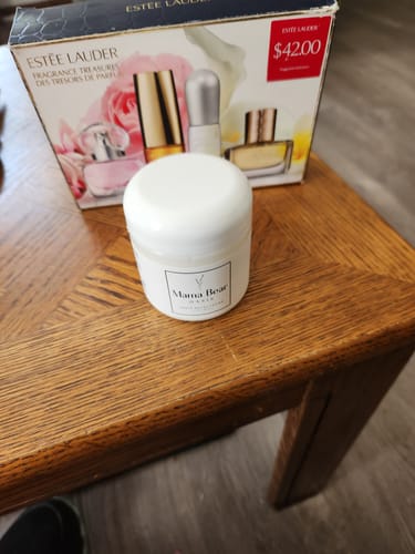 A jar of cream on a table with an Estee Lauder fragrance set in the background.