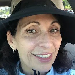 A smiling woman with dark hair and brown eyes wearing a wide-brimmed black hat, inside a car.