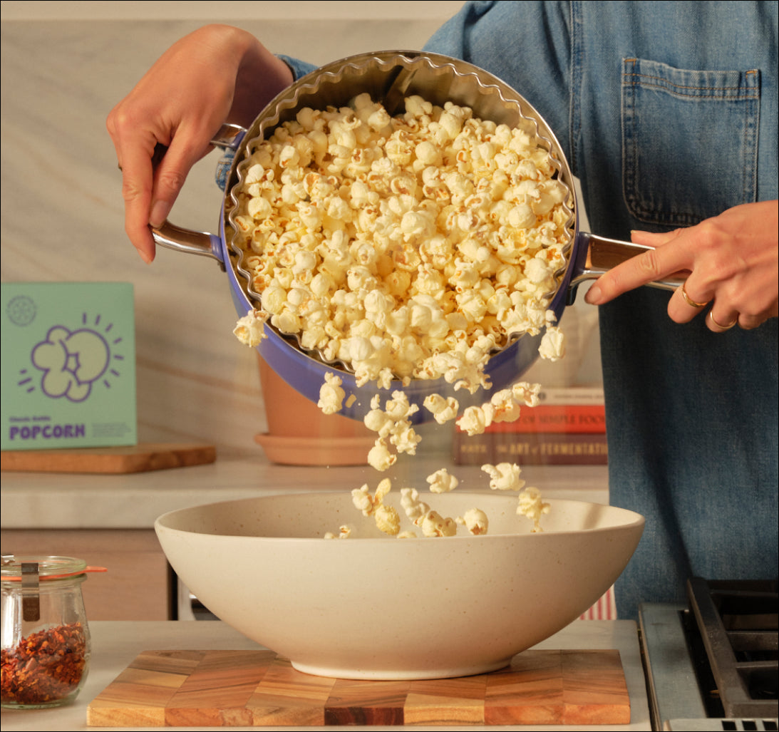 A person pours popcorn from a blue pot into a large white bowl in a kitchen setting.