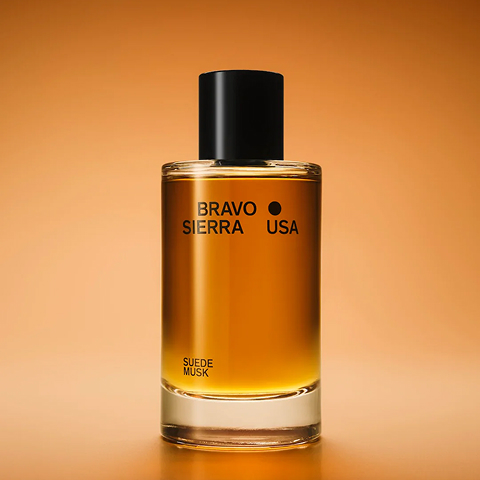 A clear glass bottle of Bravo Sierra Suede Musk cologne with a black cap on an orange background.