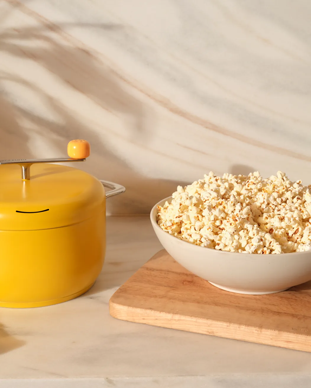 A yellow stovetop popcorn popper sits on a marble countertop next to a large bowl of popcorn.