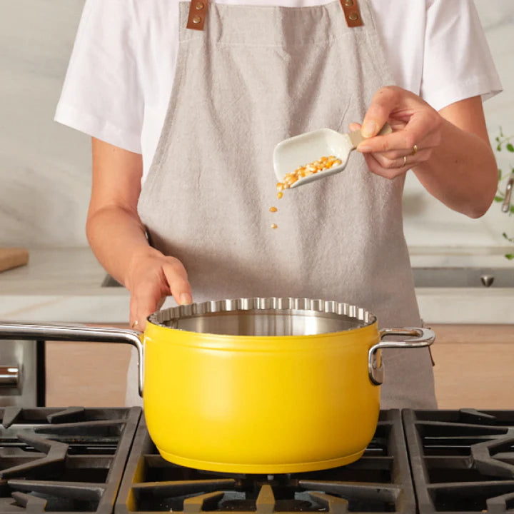 A person in an apron pours popcorn kernels from a scoop into a yellow pot on a stove.