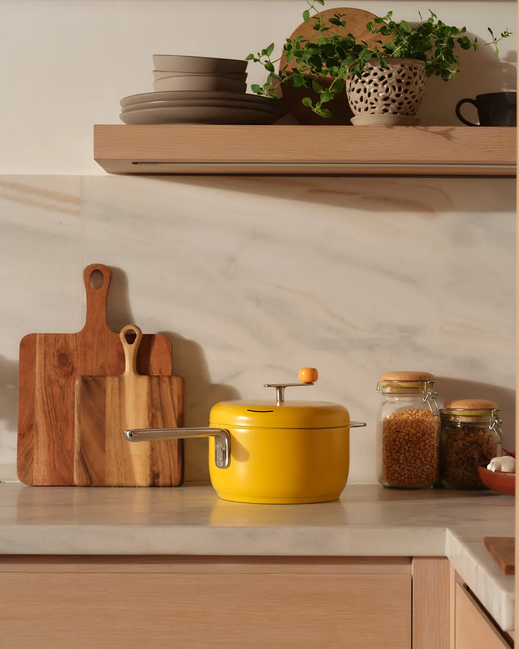 A yellow stovetop popcorn popper sits on a marble kitchen counter next to cutting boards and ingredient jars.