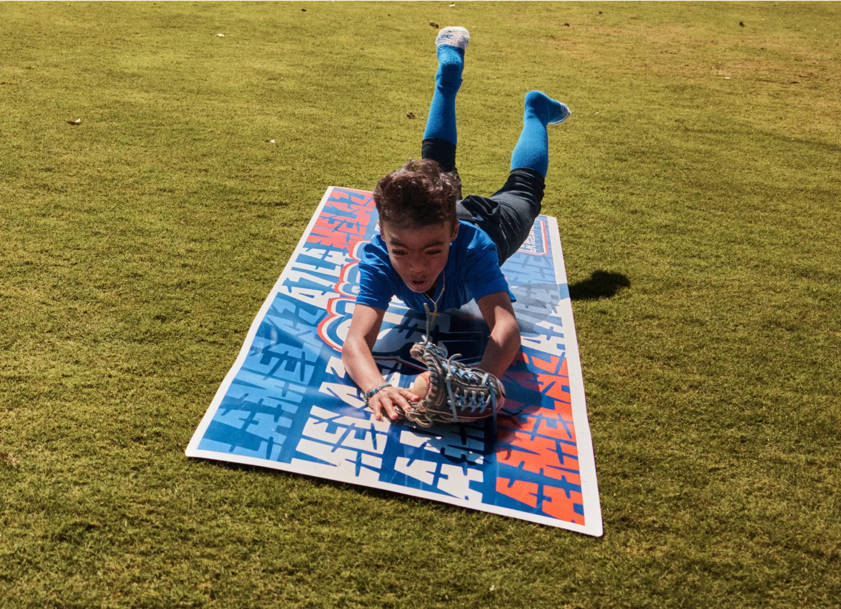 A young boy in blue lays on a colorful mat on a grassy field, reaching for a baseball glove.