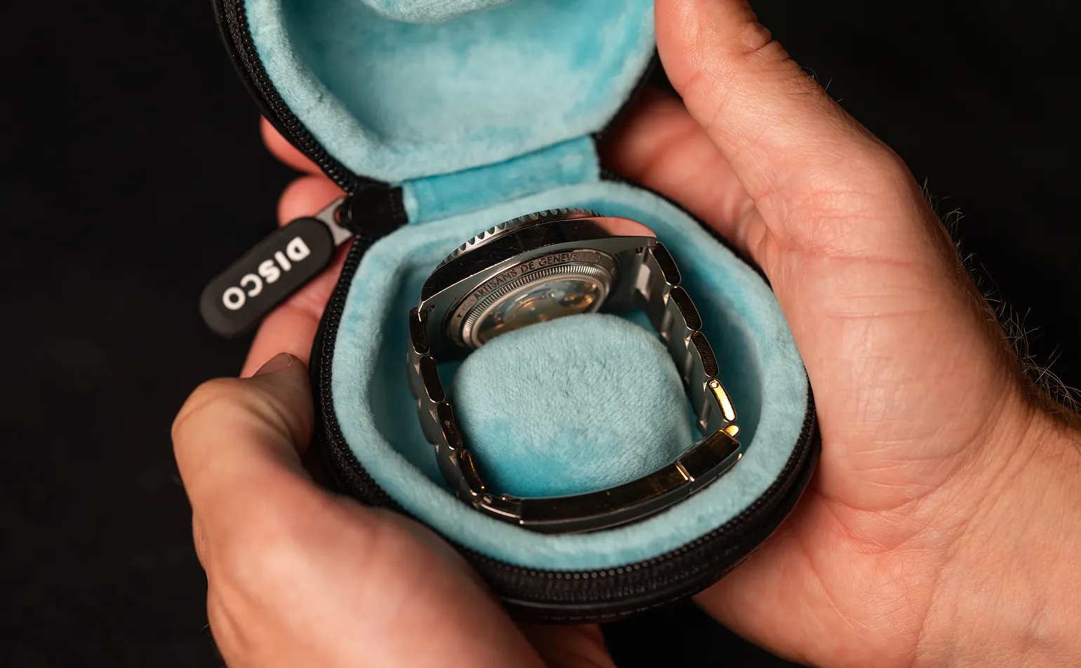 A person holds open a black zippered case with a blue interior, showing a wristwatch inside.