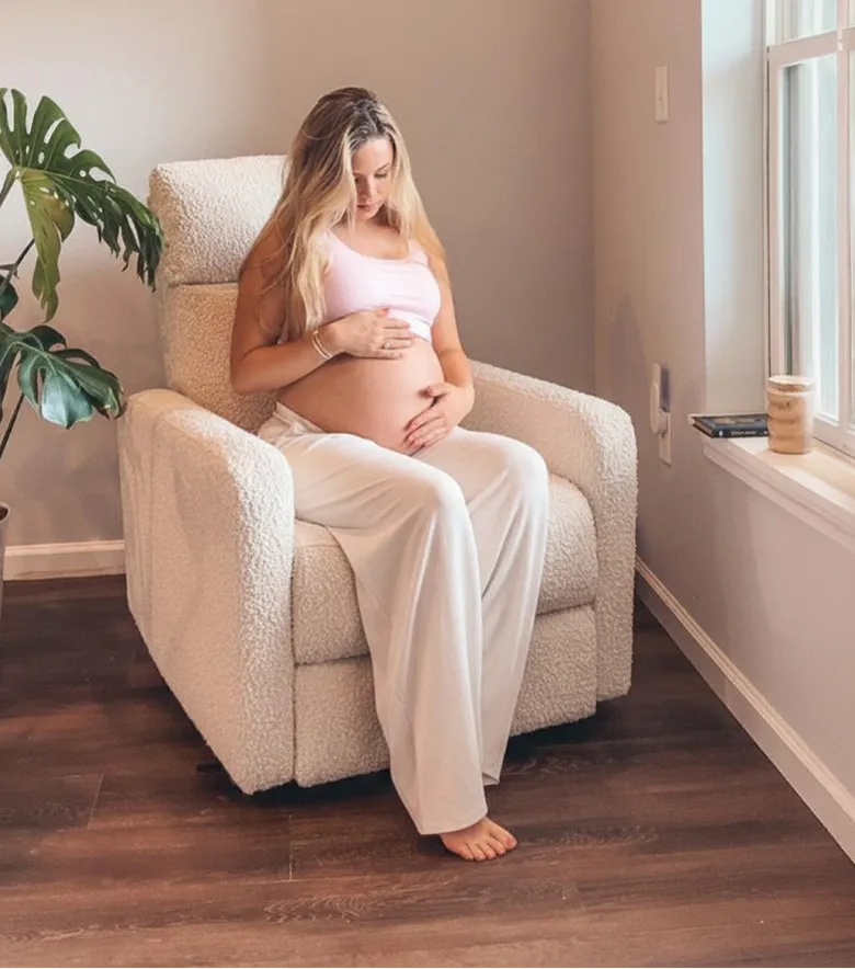 A pregnant woman in white sits in a fluffy white armchair, holding her belly and looking down.