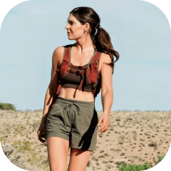 A woman in athletic gear with a hydration pack walks through an arid, hilly landscape.