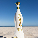 White bottle with gold lettering on a sandy beach under a clear blue sky.