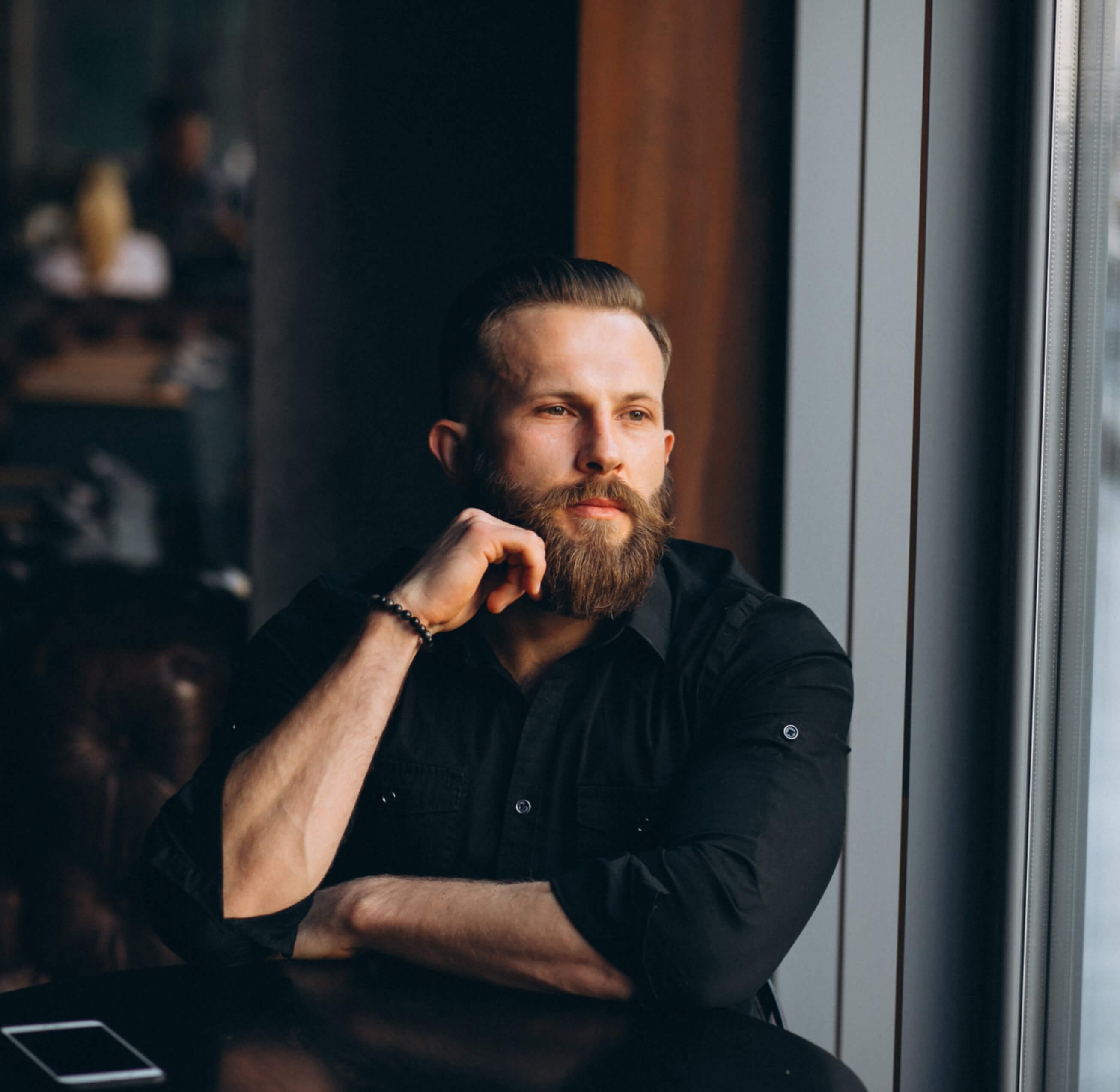 Bearded man in black shirt sitting by a window, looking thoughtful.