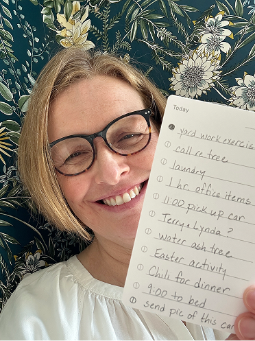 Person smiling, holding a handwritten to-do list against a floral wallpaper background.