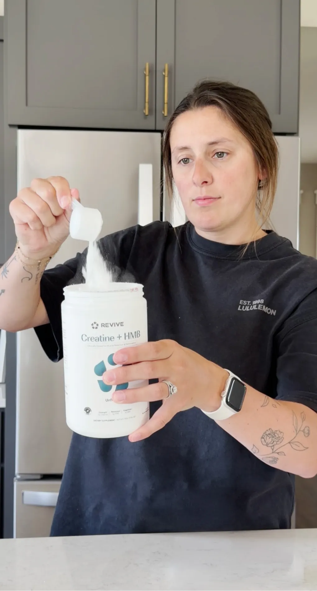 A woman in a kitchen scoops white powder from a large white tub of creatine supplement.
