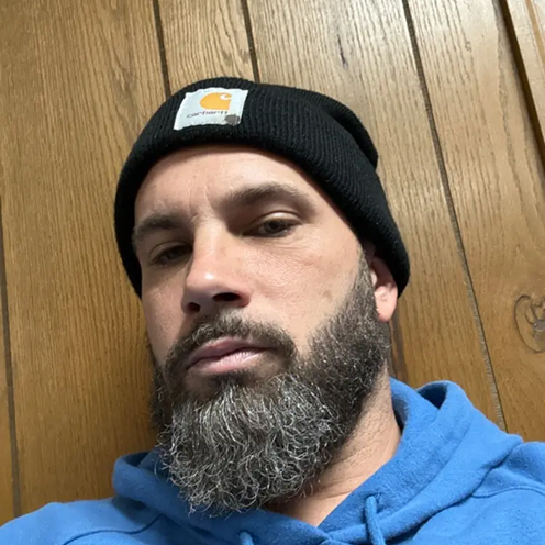 Man with a beard wearing a black beanie and blue hoodie against wooden background.