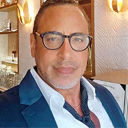 A man wearing glasses, a white collared shirt, and a blue blazer looks at the camera.
