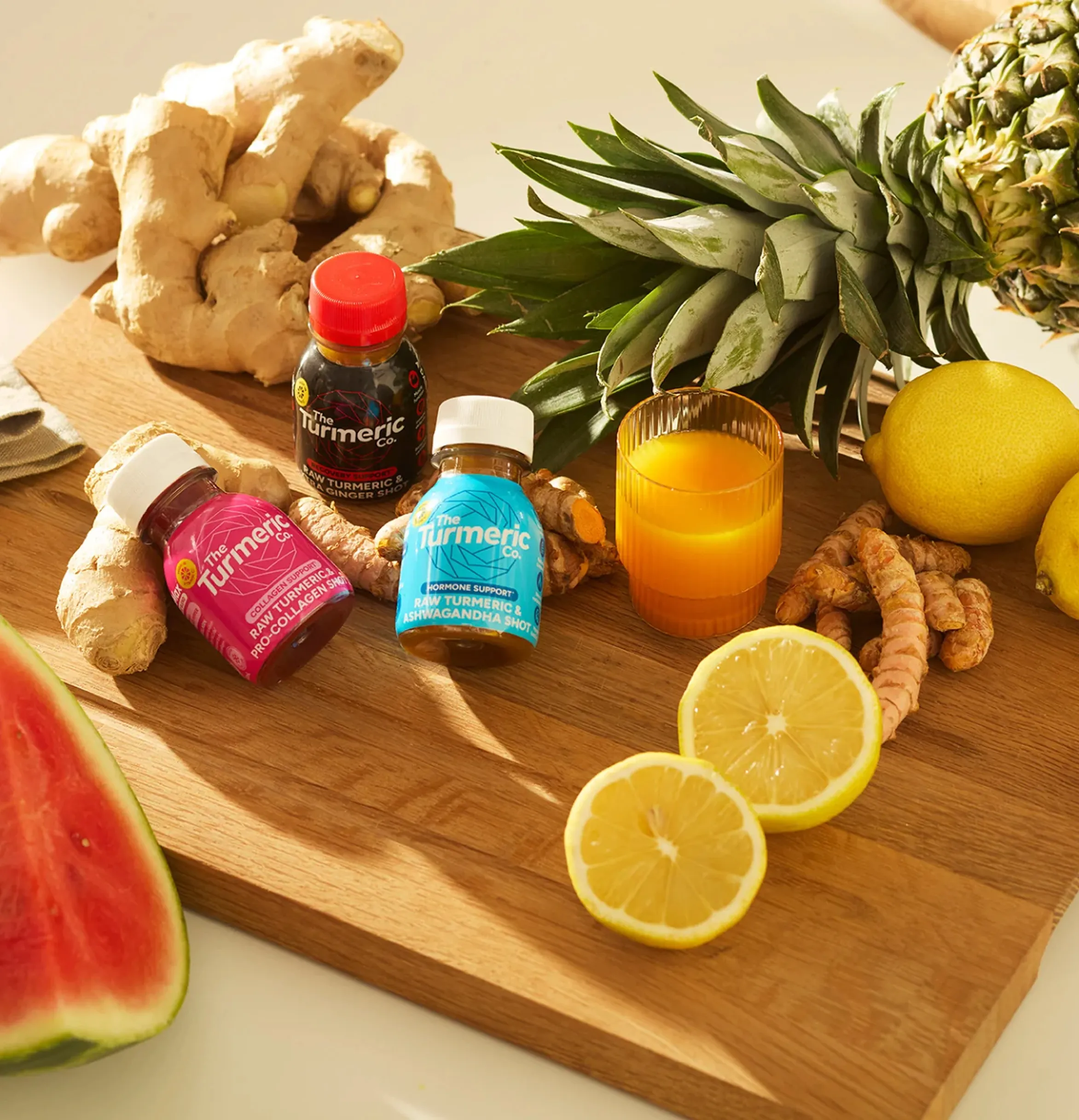 Three turmeric shots on a wooden board with fresh ginger, pineapple, lemons, turmeric root, and watermelon.