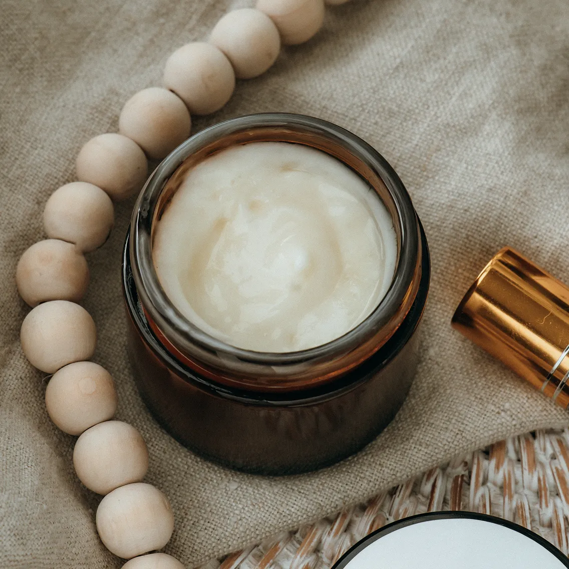 Open jar of cream beside wooden beads on fabric.