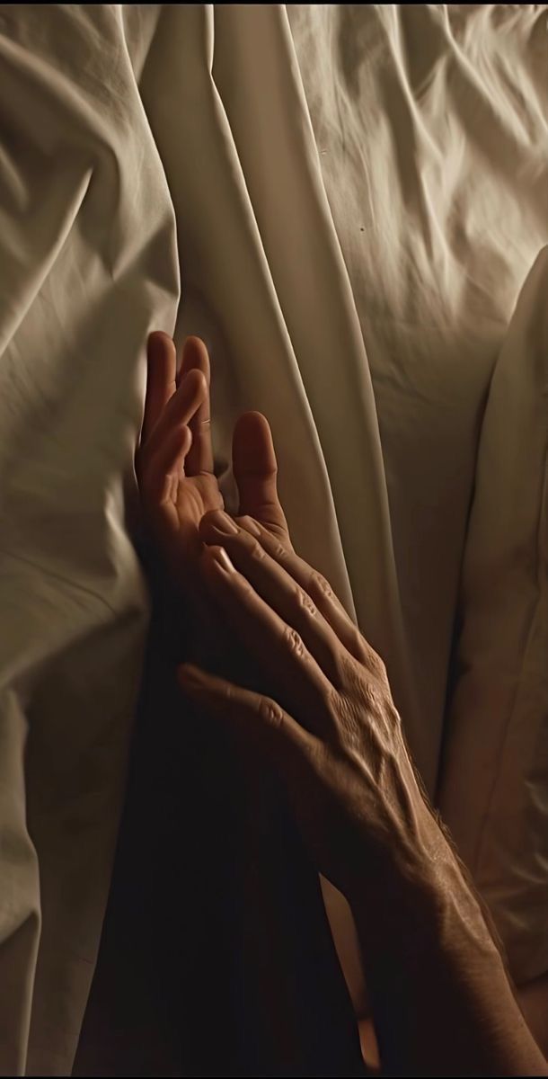 Two hands gently touching on a bed with white sheets.