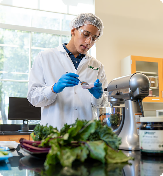 A person in a lab coat, hairnet, and goggles examines a substance in a small dish in a lab.