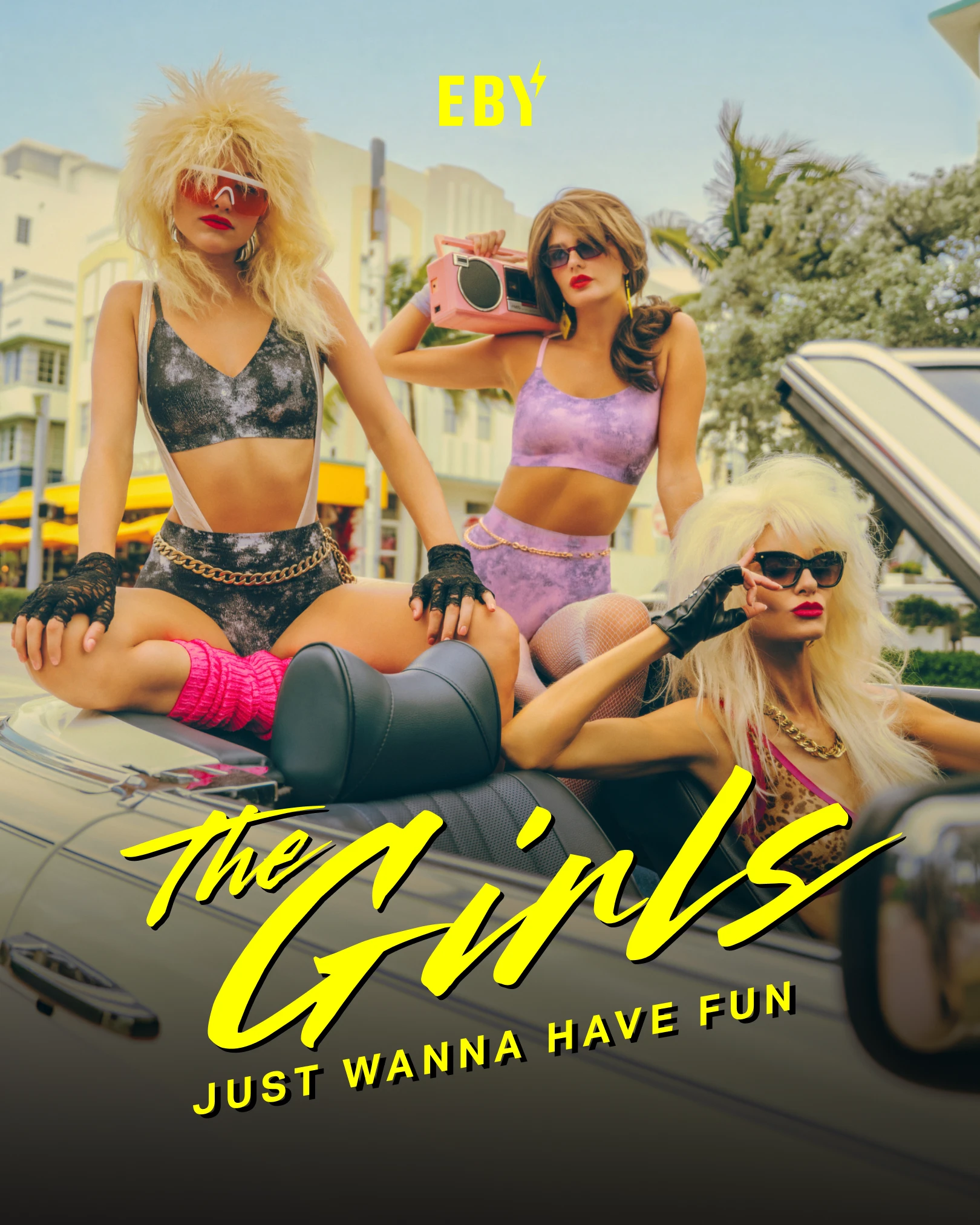 Three women in 1980s-style outfits and big hair pose on and in a convertible car.