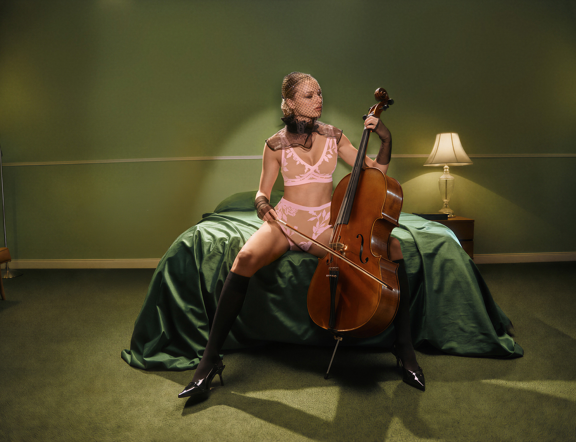 A woman in pink lingerie and a veil sits on a bed, holding a cello in a green room.