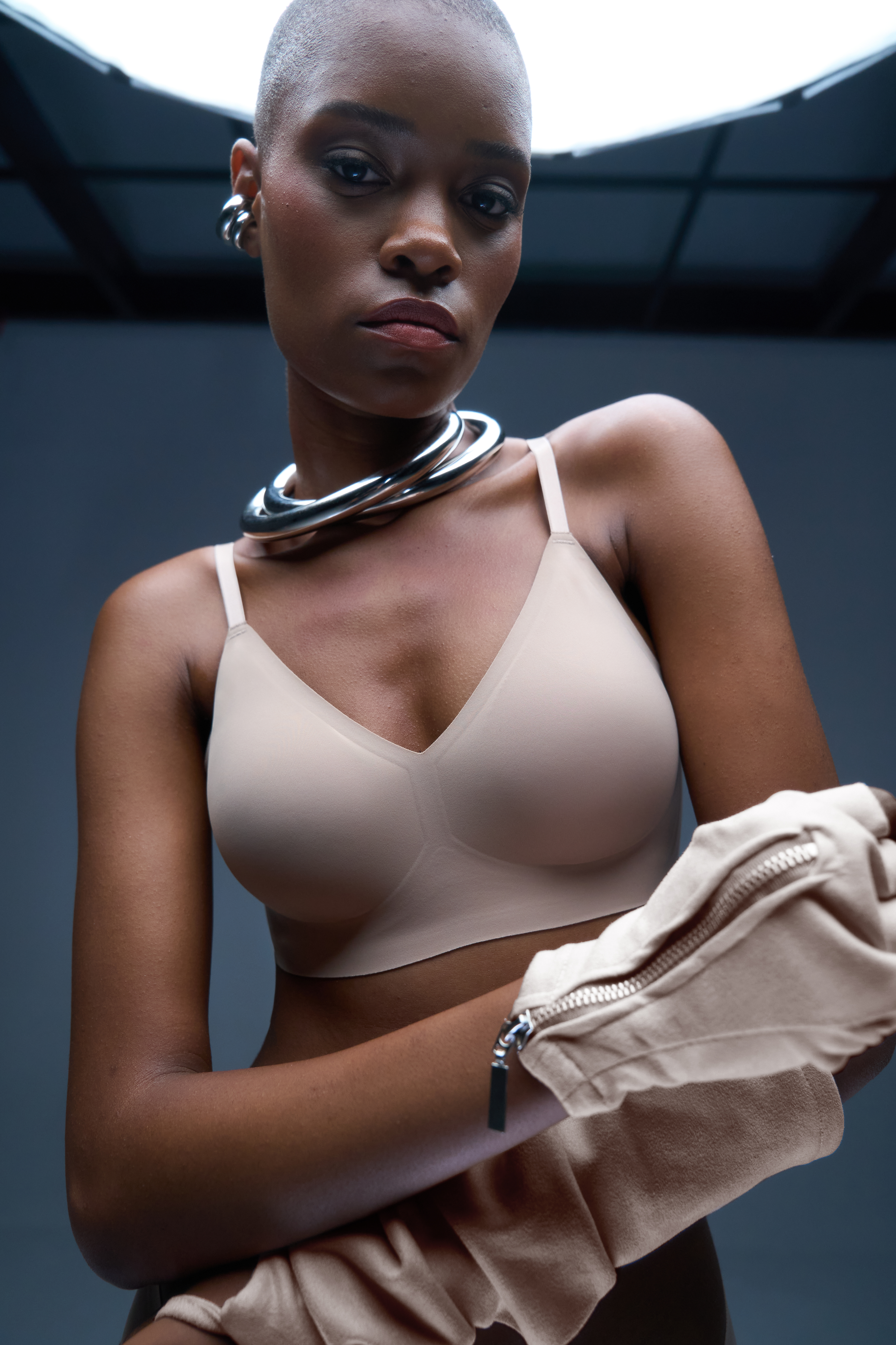 Person wearing a beige bra and metal necklace, holding beige clothing.