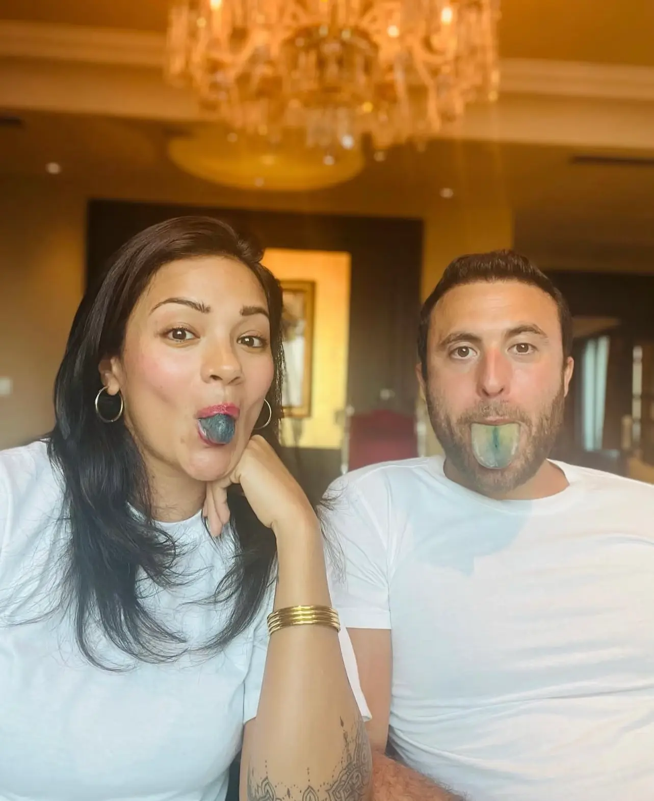Two people with colored tongues, sitting under a chandelier.