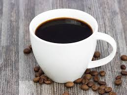 A white cup filled with black coffee, sitting on a grey wood surface with coffee beans scattered around it.