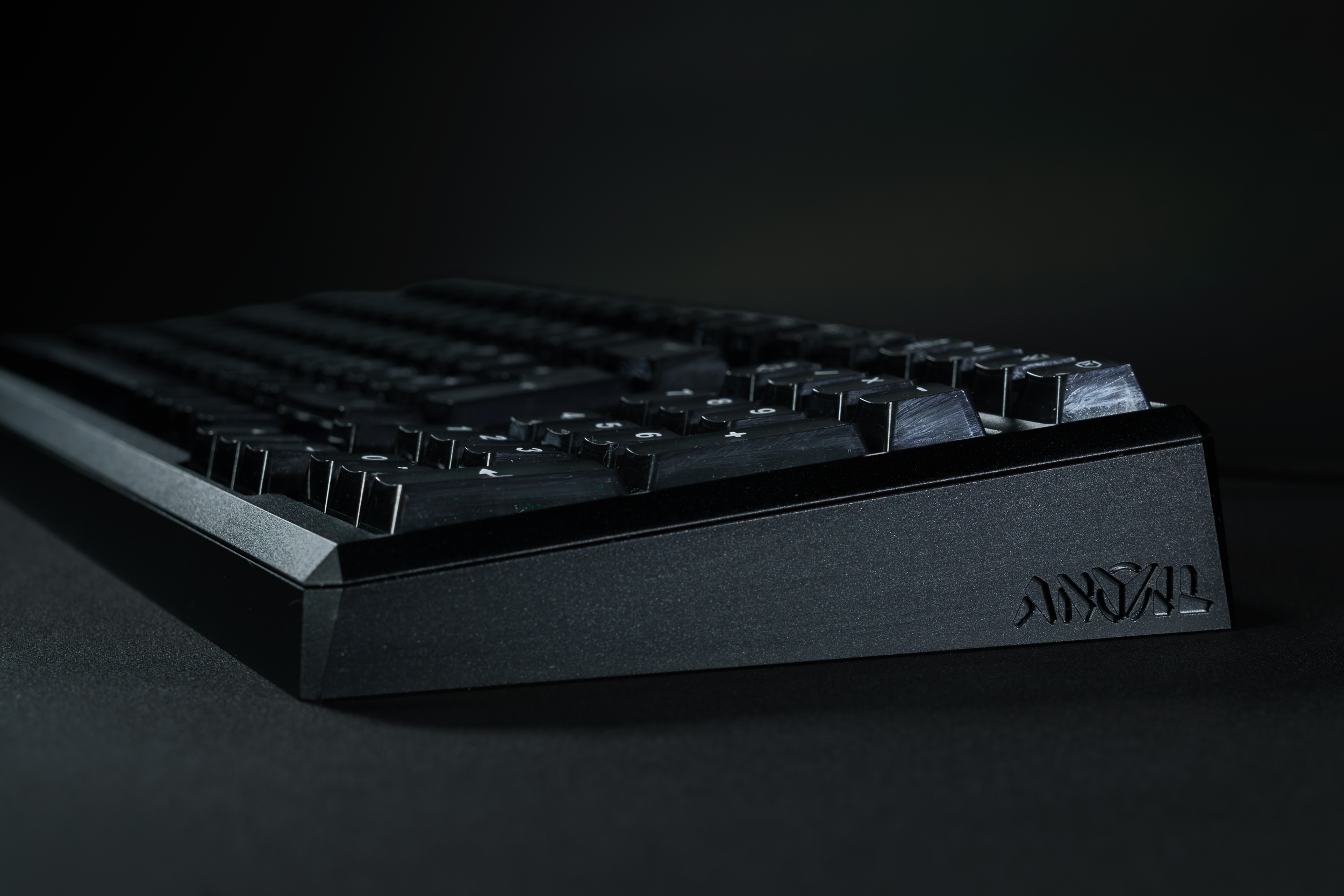 A low-angle, side profile of a black keyboard with a logo, sitting on a black surface.