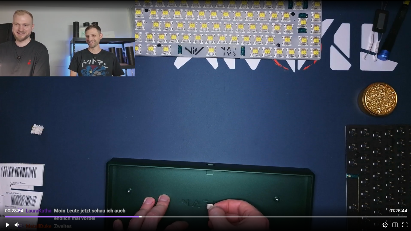 A screenshot of a video stream where two men watch hands assemble a custom mechanical keyboard on a desk.
