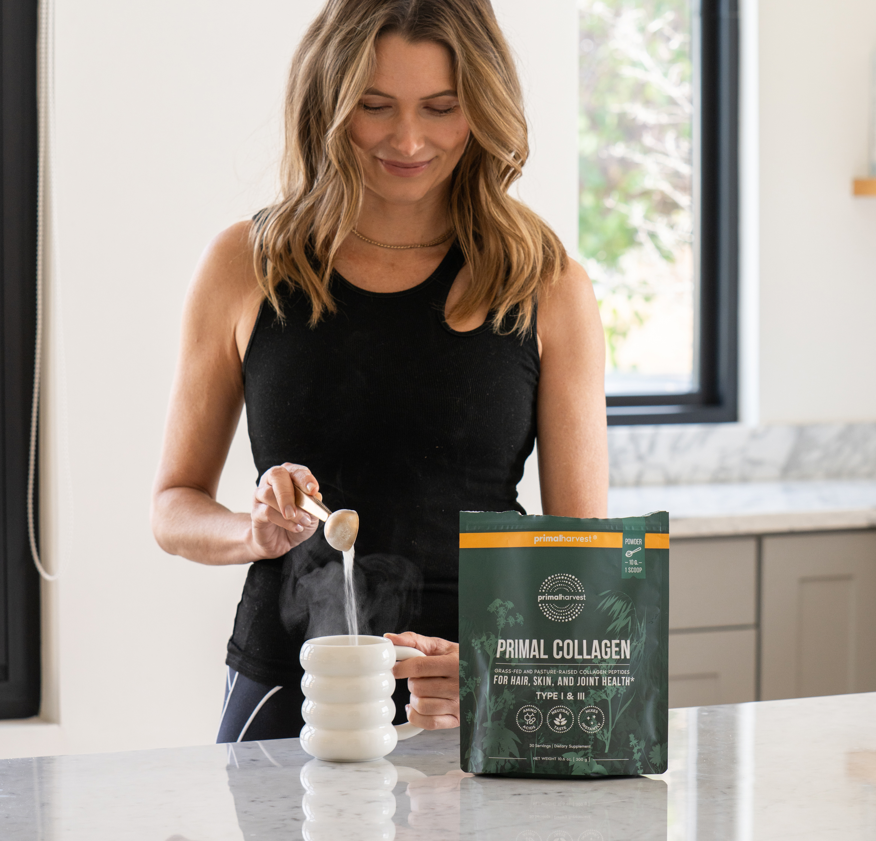 Woman adding Primal Collagen powder to a steaming mug in a kitchen.