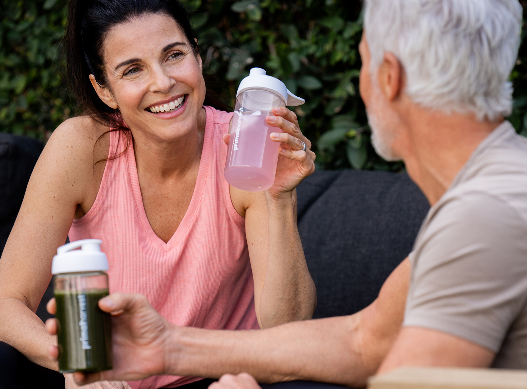 Two people sitting outdoors, holding bottles with drinks, smiling at each other.