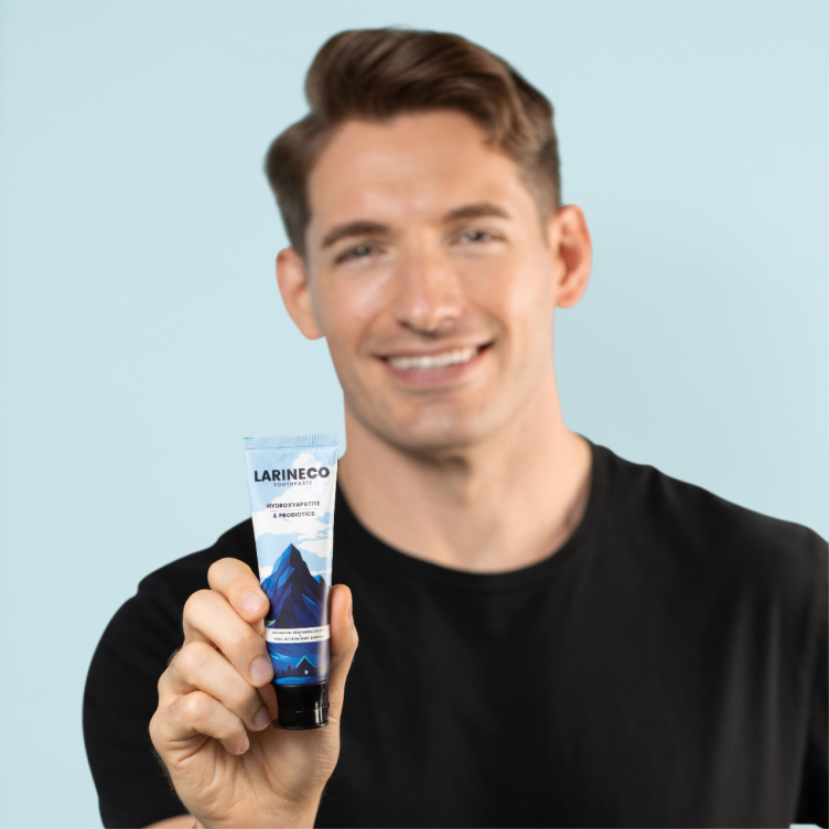 A smiling man in a black t-shirt holds up a tube of Larineco toothpaste against a blue background.
