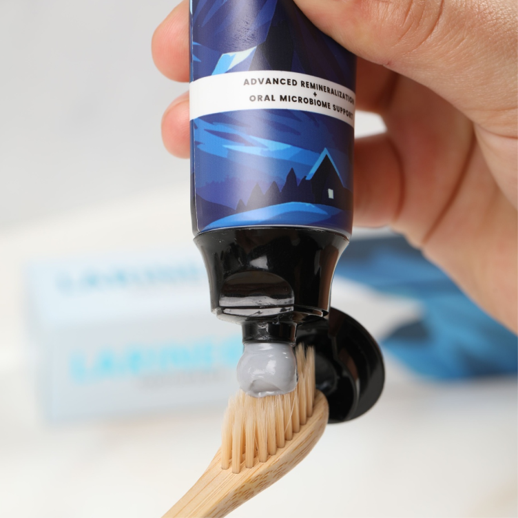 A hand squeezes grey toothpaste from a blue and black tube onto a wooden toothbrush.