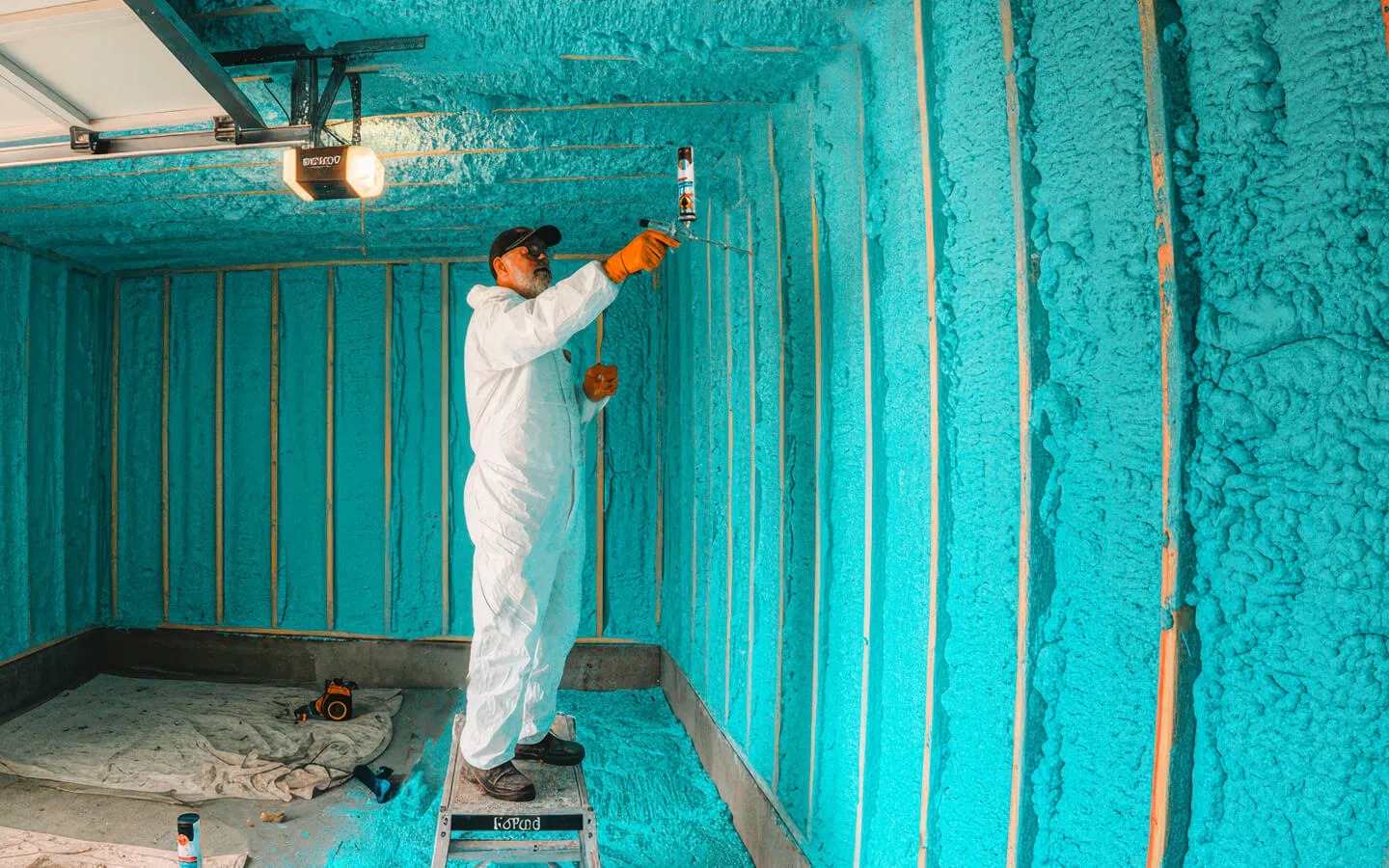 A worker in a white protective suit applies bright blue spray foam insulation to garage walls.