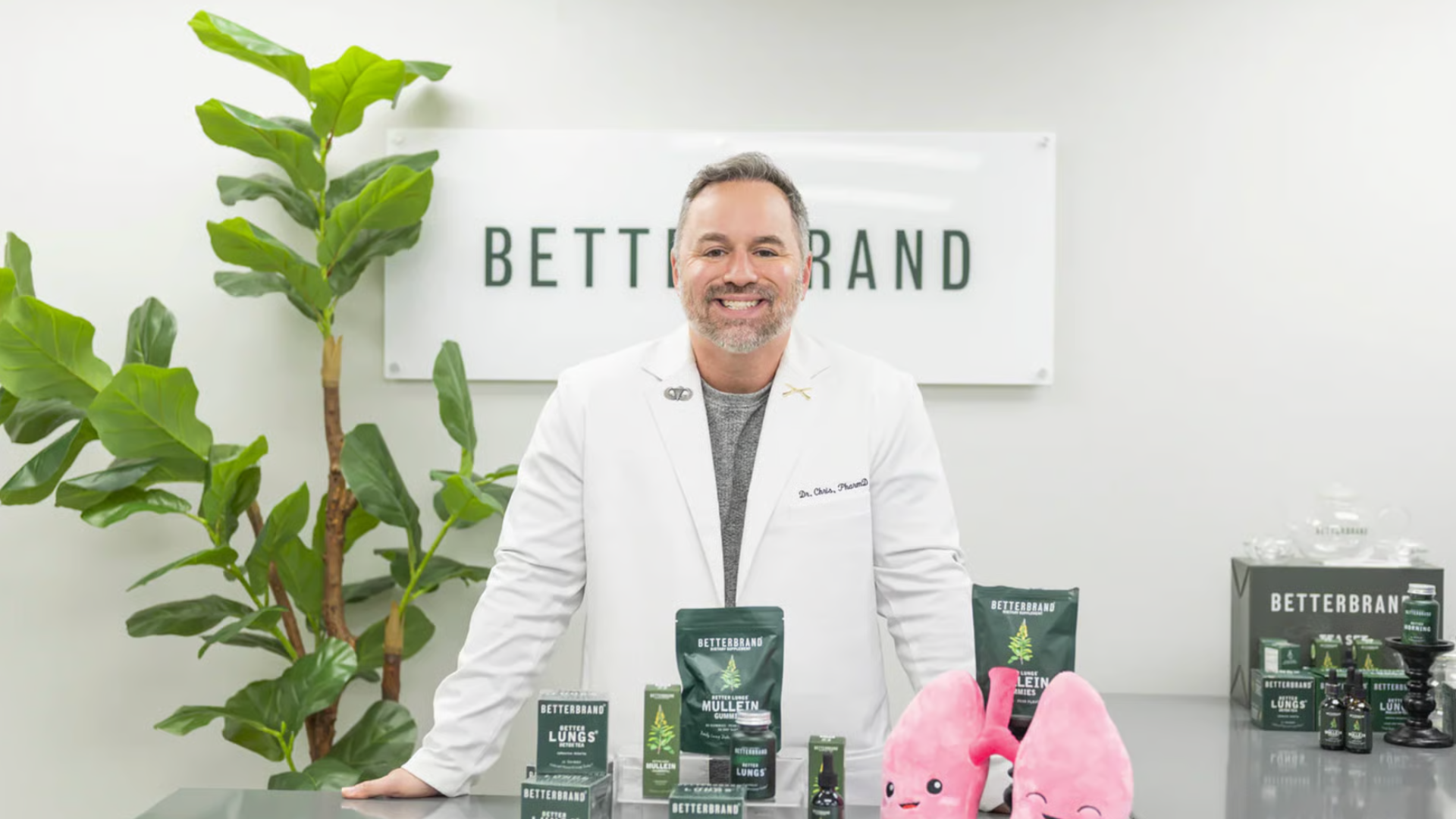 Man in a white coat smiles behind health products and plush lungs on a table.