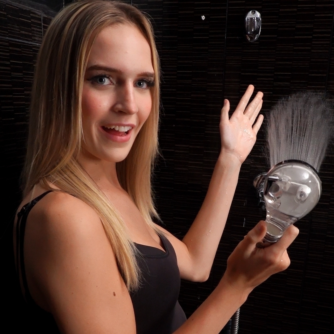 Person holding a showerhead with water spraying, smiling in a bathroom.