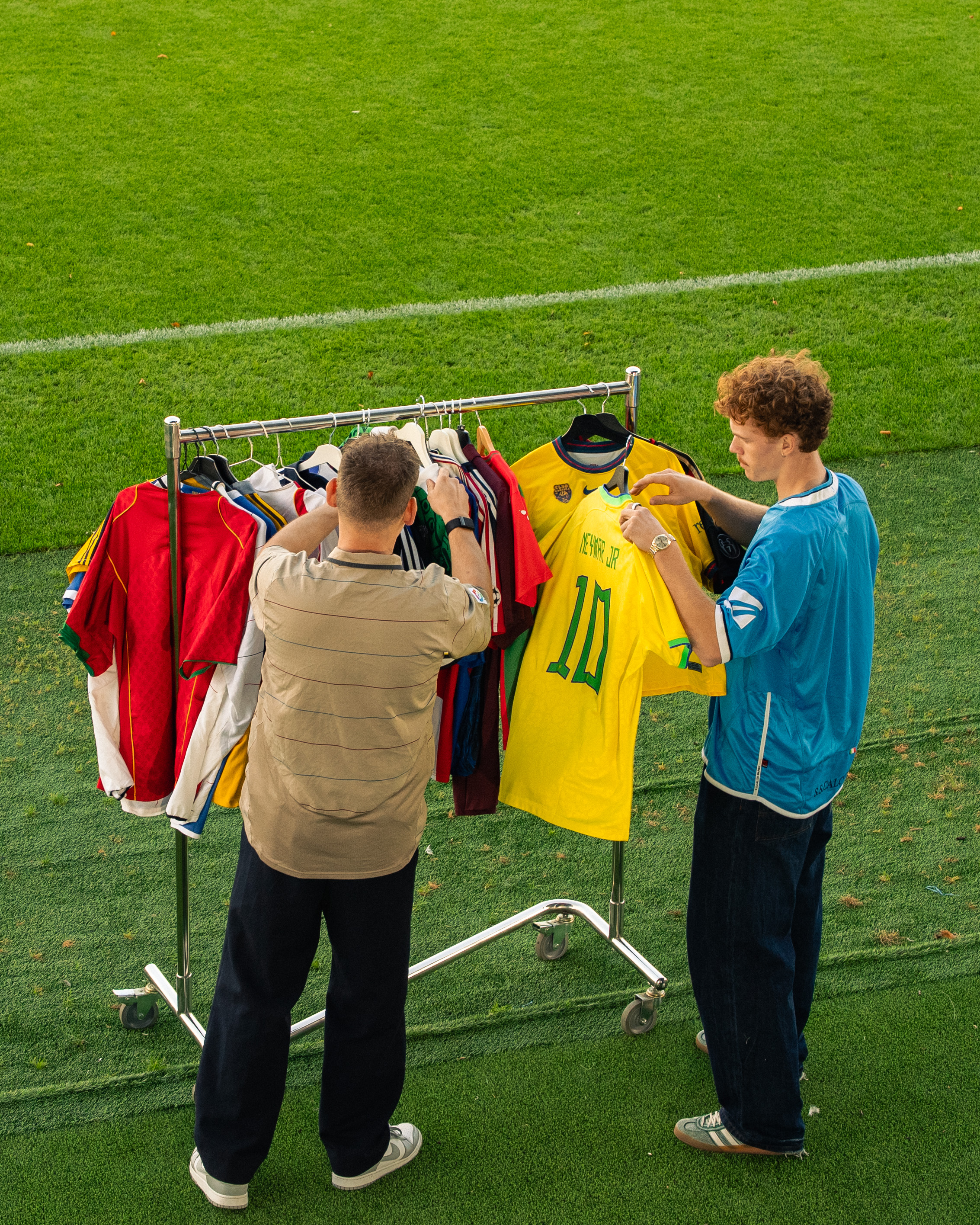 Two people browsing soccer jerseys on a rack by a grassy field.